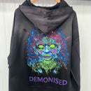 Black hoodie with a colorful skull design and 'Demonised' text on the back, hanging against a white wall.