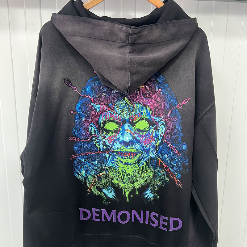 Black hoodie with a colorful skull design and 'Demonised' text on the back, hanging against a white wall.